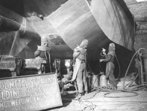 Black and white photograph of three people spray painting the underside of a boat