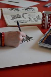 Close up of a person drawing a print of a mythical creature. Their hand and the drawing is in view, accompanied by sketchbooks and coloured artist pencils.