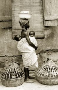 Photograph of a Yoruba woman with two babies in her arms and carrying baskets on her head