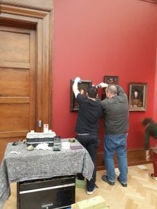 Two technicians work on hanging other paintings in the gallery. One of them holds the painting while the other uses a drill to attach it to the wall.