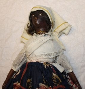 A close up of one of the dolls. There's white cloth wrapped around the head with a gold hem, a gold necklace, a white cloth top and a dark blue floral skirt. 