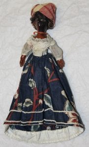A picture showing the whole entirety of one of the dolls. There's red and white cloth wrapped around the head, a red necklace, a white cloth top and a dark blue floral skirt. 