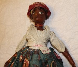 A close up of one of the dolls. There's red cloth wrapped around the head , a white cloth top, a red necklace and a blue floral skirt. 'MM' is also embroidered on the pocket