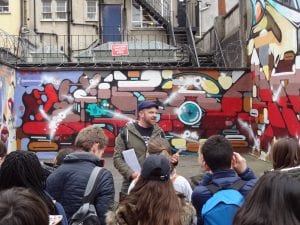 Rob Wheeler stood in front of a graffitied wall explaining it's significance to a group of walkers