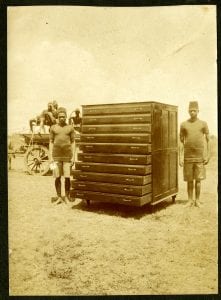 Photograph of two Kenyan apprentice carpenters standing either side of a large chest of drawers.