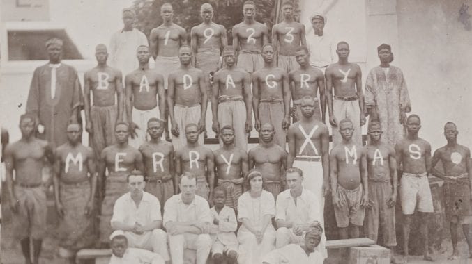 Men in rows are standing with text written on their bodies. "1923 Badagry Merry Xmas." A white family are sitting at the front. Featured image in Empire Through the Lens