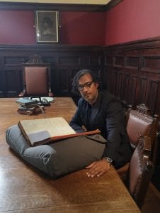 David Olusoga on the set of 'A House Through Time'. He's sat in front of a large archive casebook that's resting on a cushion.