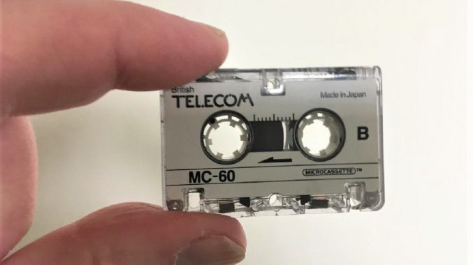 A microcassette (tiny cassette tape) held between a thumb and forefinger to illustrate its small size