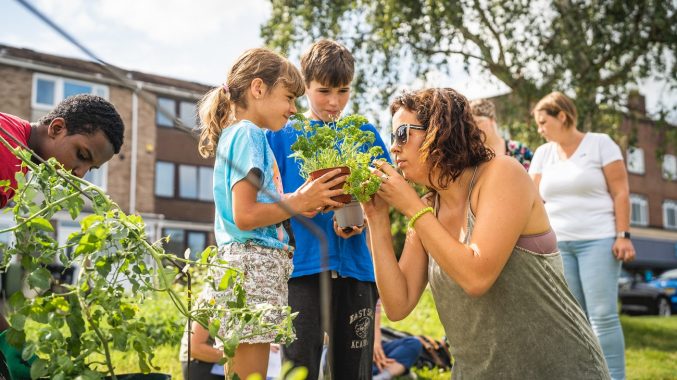 A boy and girl hold a pot of parsley for a woman to smell. An older boy to the left tends plants, and two women are talking in the background. It's a sunny day in a green space near some houses.