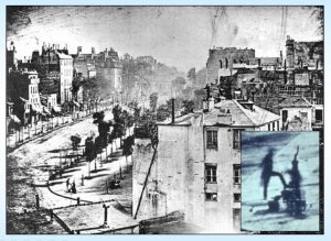 Photograph of first human subject taken by Louis Daguerre. View of the Boulevard du Temple in Paris, 1838. There are trees lining the street and there is a silhouette of a man getting his shoe shined. Either side of the street are lined with buildings. 