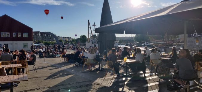 People sat in the sunshine at M Shed cafe with hot air balloons rising in the distance