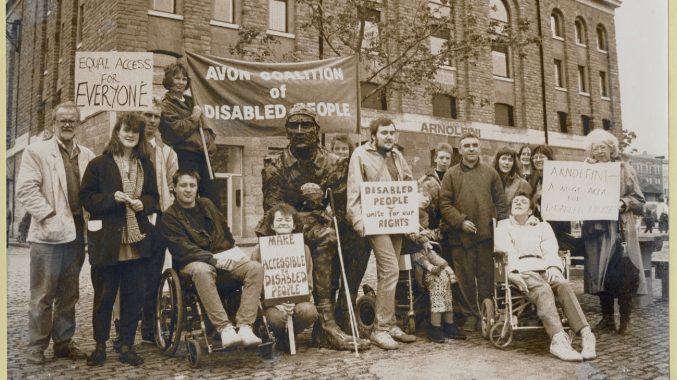 Black and white photograph of the Avon coalition of disabled people. There is a group of people with placards