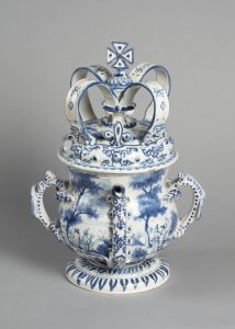 Photograph of pot with four handles going all the way around. It has grand ornate lid, similar shape to a crown. There is paintings in blue all around the circumference of the pot depicting trees. The rims of the handles and base are lined of have a dotted pattern.