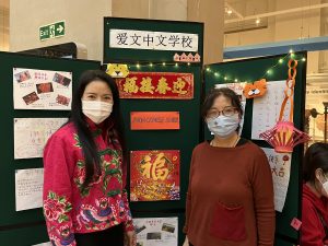 Two women with face masks on stood in front of a stand promoting Chinese activities