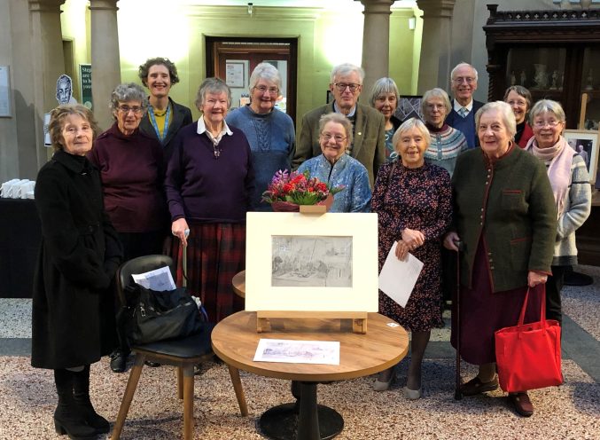 Twelve older people stood in the rear hall at Bristol Museum behind a drawing by Muller. Curators Kate Newnham and Karin Walton are stood with the group.
