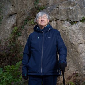 A portrait of an older woman stood holding a walking stick against a rocky background