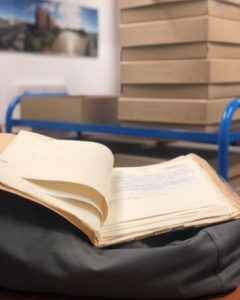 An open book lying on top of a blue cart in a British Empire archive room. There are cardboard boxes in the background.