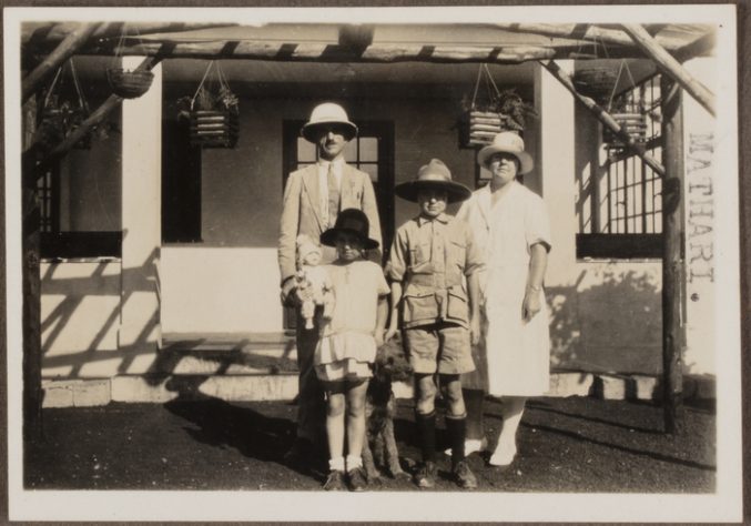 Black and white photo of a family of four stood in front of a house, all wearing hats. It's a sunny day.