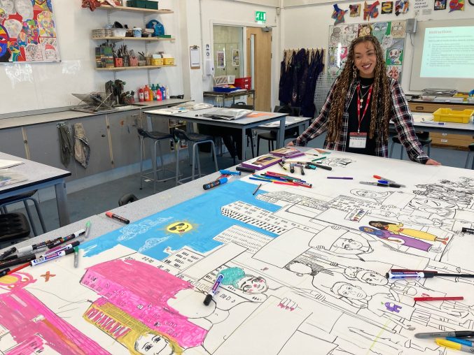 Jasmine Thompson looking over a large partly coloured in mural on the table. She's standing in a classroom full of art supplies and an interactive whiteboard is on the wall behind her. Coloured pens are scattered across the table in front of her.