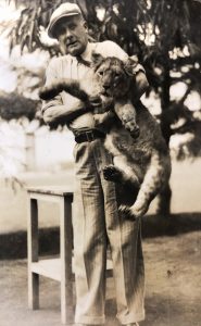 Black and white photo of a man holding a lion. He's wearing a cap and there's a tree in the background.