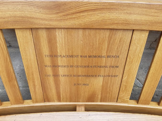 A close up of an oak bench with an inscription that reads 'This replacement war memorial bench was provided by generous funding from the Post Office Remembrance Fellowship, June 2022 