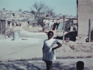 A street scene in Southern Africa showing a young Black woman looking at the camera, filmed by Theresa Knowles, c. 1956