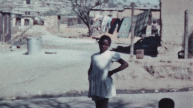 A street scene in Southern Africa showing a young Black woman looking at the camera, filmed by Theresa Knowles, c. 1956