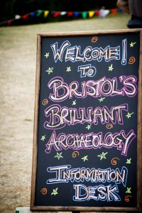 Black a-board reads "welcome to Bristol's brilliant archaeology - information desk" in colourful chalk