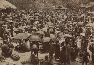 Crowd of people, some holding umbrellas, at a market.