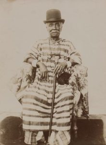 Man with a moustache sat wearing a long, stripped gown, a dark top hat and leaning on a cane.