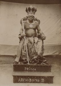 Topless man sat on a throne which reads 'Prince Archibong II'. He wears a crown and holds a sceptre.