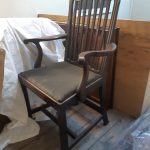 Shows a huge wooden chair facing to the left. Behind it are dust sheets which were used to protect the chair from dirt. The chair has a high back with slats, a lightly padded dark seat, and long wooden armrests.