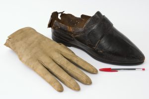 These are O'Briens shoes and gloves. They are very large, and the glove is in front with the shoe behind it. The glove is pale yellowish cream and is made of a soft thick animal skin, possibly suede. The shoe is very dark leather, wit ha strap across the front. It has probably been worn, as there is a small tear at the back.