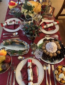 Festive Victorian table set up includes decorative plates laid with crackers, cutlery and red napkins, glassware, silver candle holder as well a turkey, fruit bowl and a plate of chestnuts arranged nicely.