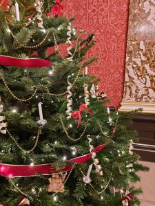 Details of Christmas tree decked with lights, red ribbon, curled gold paper, gold beads and candles. Patterned red wallpaper in the background.