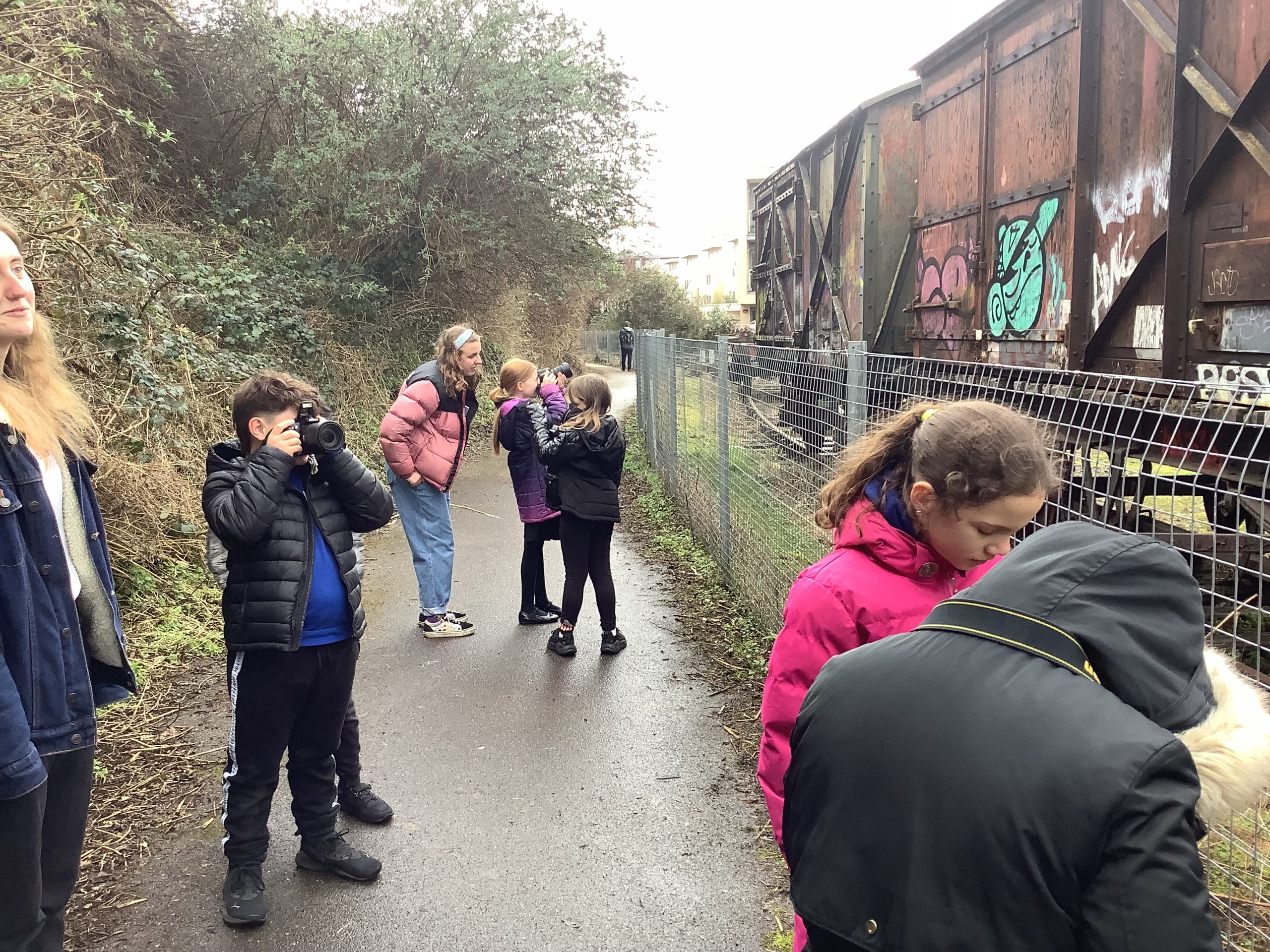 group of primary aged children using professional cameras. 2 adults assisting, on a path beside a railway carriage on Bristol harbourside