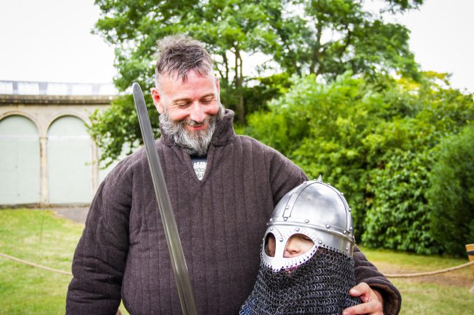 Smiling man, dressed in medieval outfit standing next to youn boy in a medal medieval soldiers helmet, carrying a sword. Part of summer activities at Blaise Museum