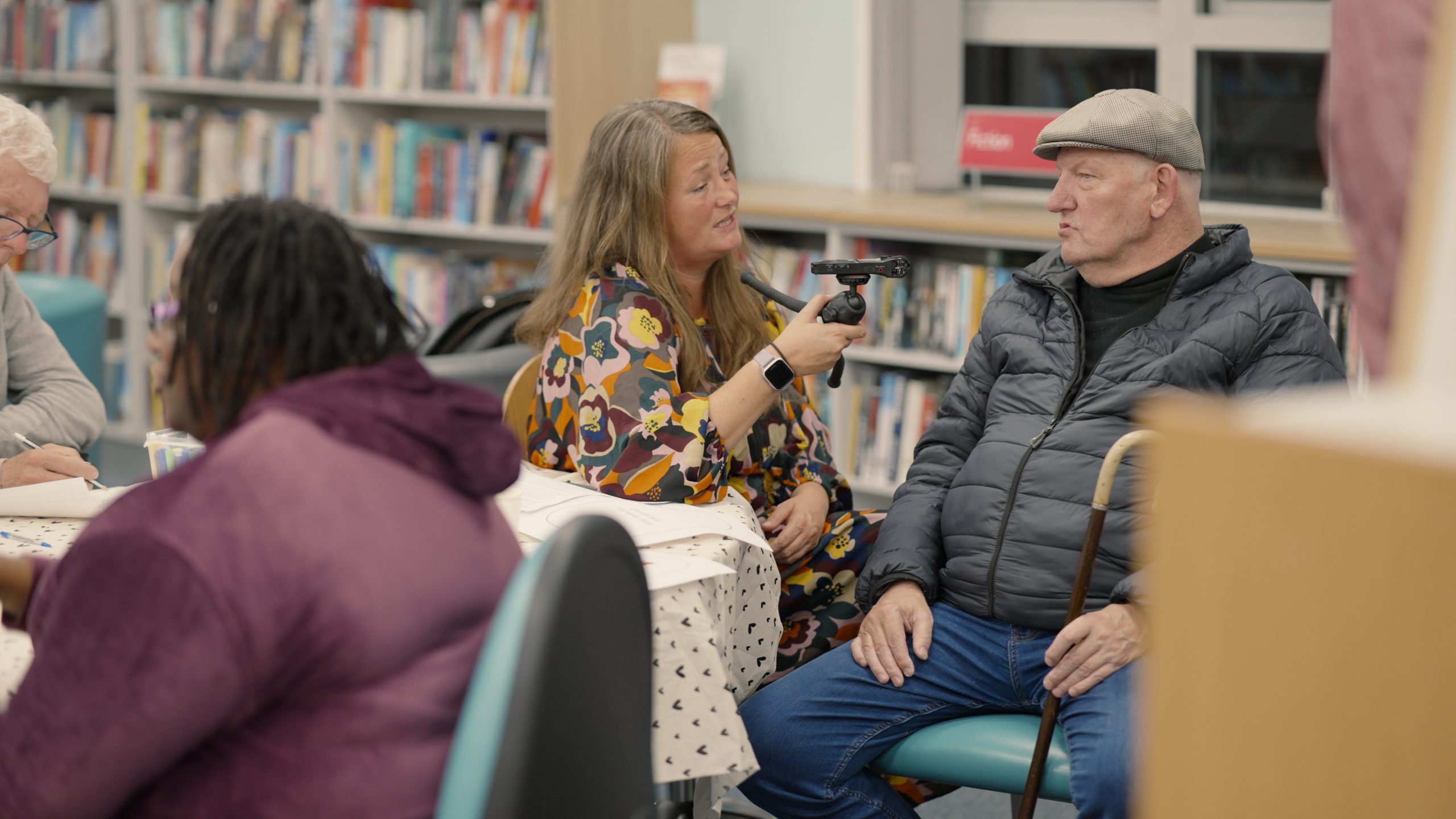 People sitting in a library. A middle aged woman hold a microphone and recording device up to an old man who is speaking into it.