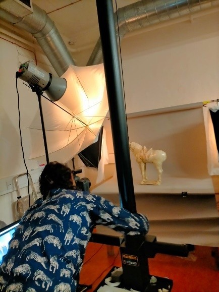 A woman with dark hair in a blue shirt taking a photo of a horse statue with a large light diffuser to her left.