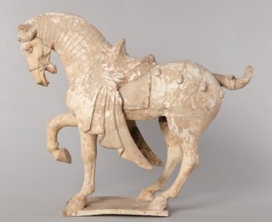 A statue of a horse wearing a saddle, with one front leg raised. The statue is made of a pale stone like material. 