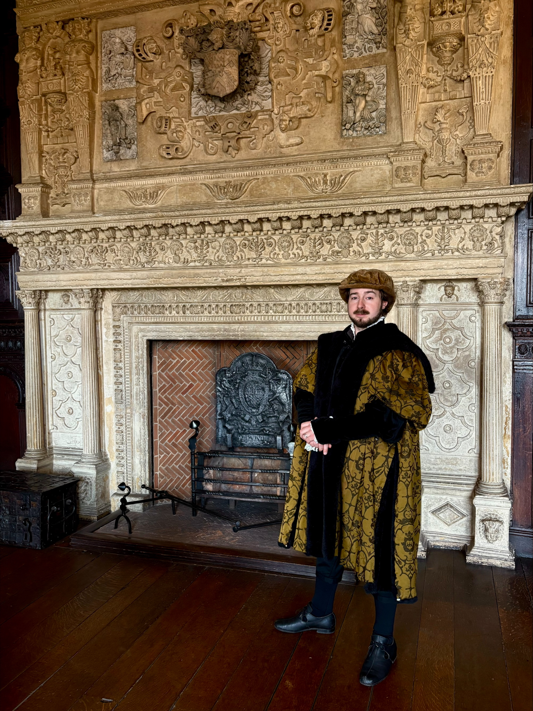 a man in Tudor costume stands beside a ornate stone fireplace in a great hall