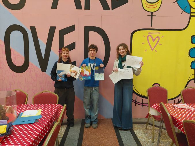 three students stand by a colourful background displaying some designs they have created