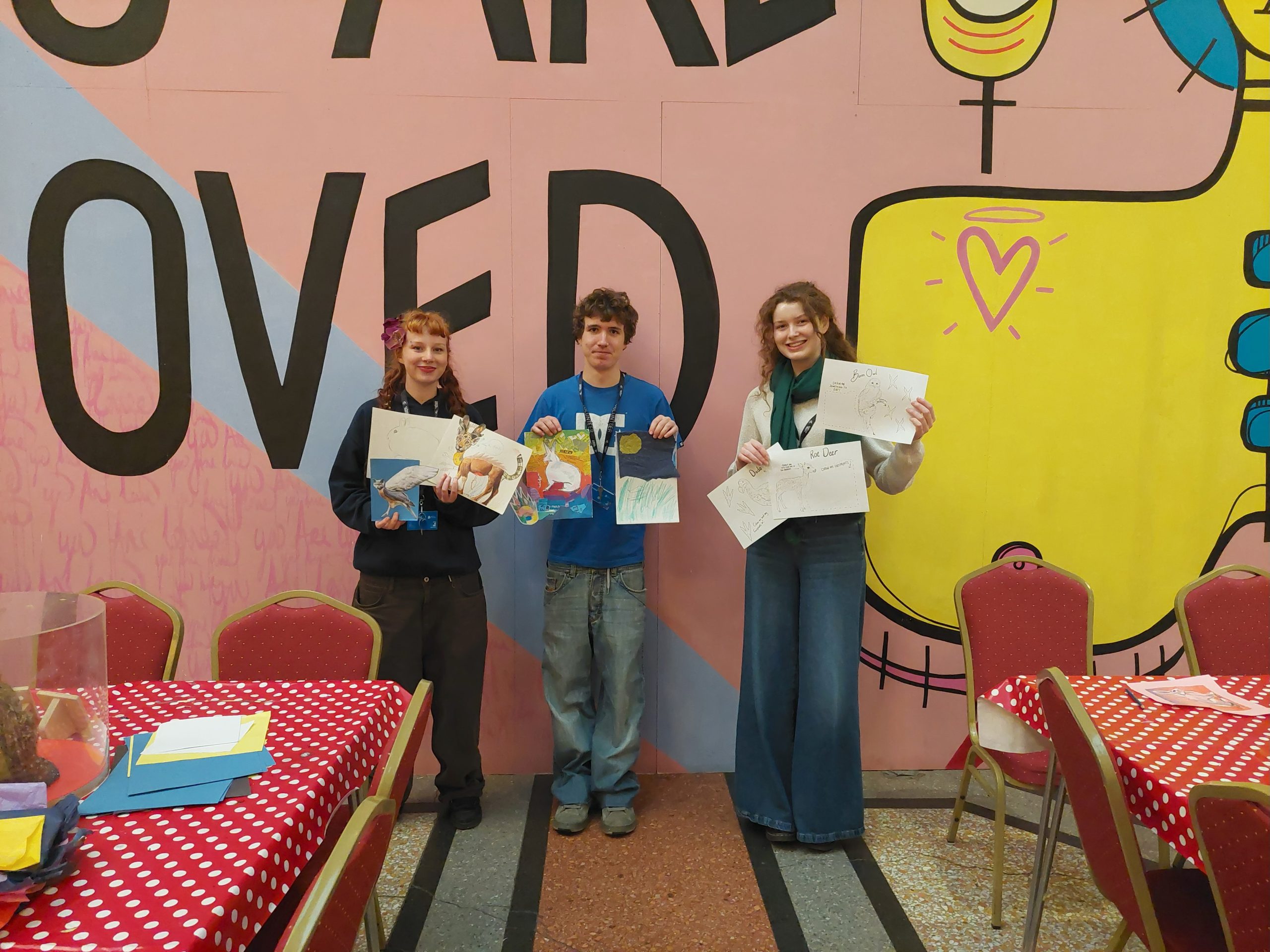 three students stand by a colourful background displaying some designs they have created