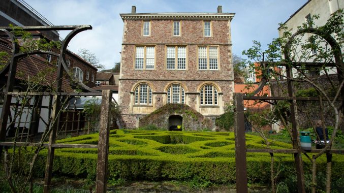 A large rectangular red brick house behind a hedged garden and iron wrought gates
