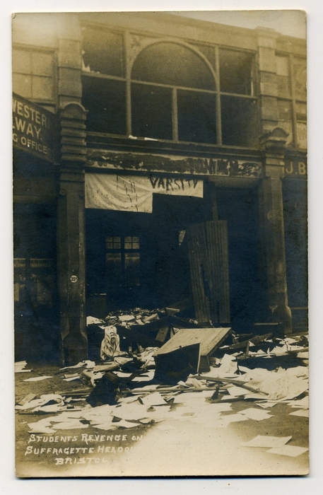 Postcard showing the headquarters of the Suffragettes, on Queen's Road, following the vandalism done by the University students. "Varsity" is scrawled above the entranceway, with wrecked furniture and papers strewn across the pavement. 