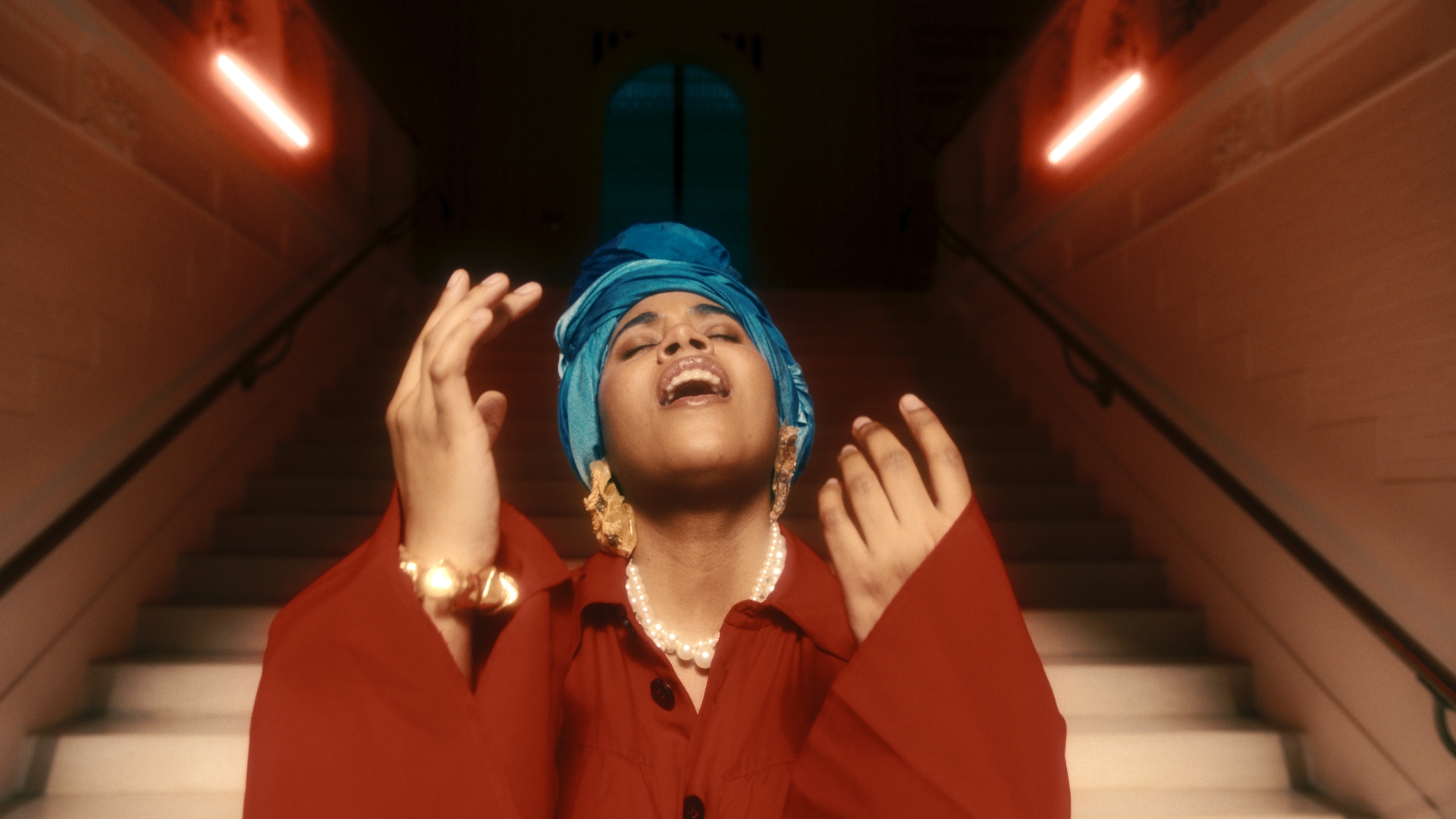 A black woman in a blue headscarf singing with her eyes closed and hands raised